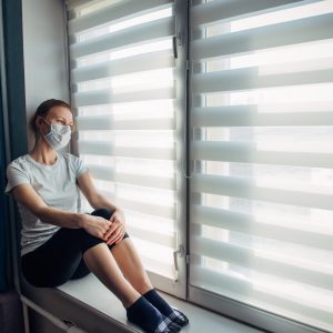 Side view young woman in wearing protective face mask sitting on windowsill at home, looking outside. Self-isolation, quarantine coronavirus pandemic prevention. Epidemic and prevention concept.